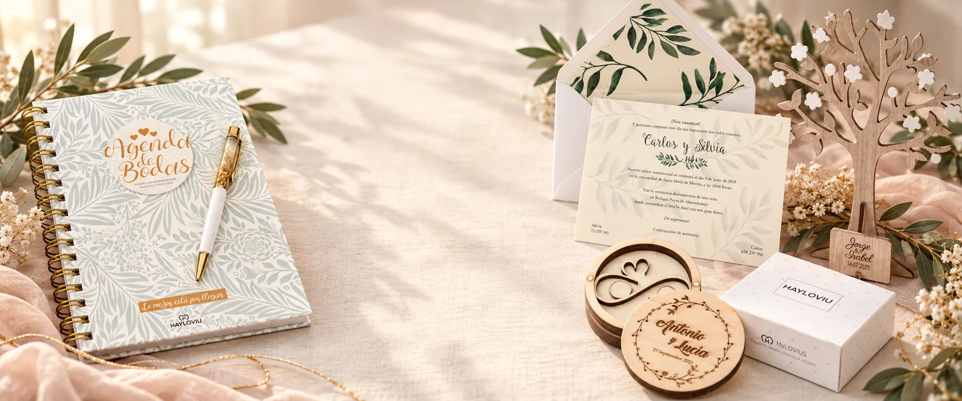 Agenda de bodas, invitación con sobre, porta alianzas grabado y árbol de deseos de HAYLOVIU sobre una mesa de lino con ramas de olivo